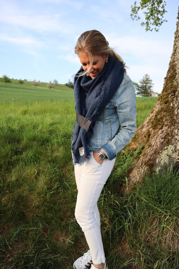 Blonde Frau in weisser Hose mit Jeansjacke und blauem Schal steht auf einer Wiese vor einem Baum.
